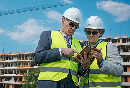 Two Oktopaz engineers wearing personal protective equipment study a project on a tablet in front of a tower crane and a building under construction - thumbnail