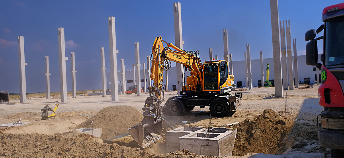 An excavator backfills by foundation footings in front of a series of mounted columns on the CTP Kac industrial facility construction site, where Oktopaz renders construction supervision - banner