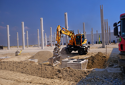 An excavator backfills by foundation footings in front of a series of mounted columns on the CTP Kac industrial facility construction site, where Oktopaz renders construction supervision - thumbnail